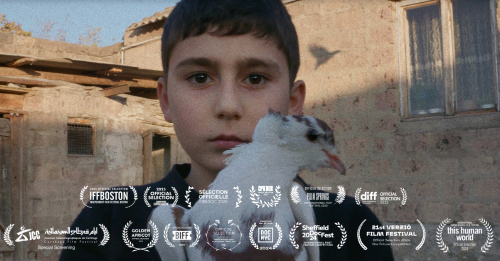 A solemn Armenian boy holds a white dove in front of a stone house in Artsakh, from the film My Sweet Land, a powerful story of war and exile.