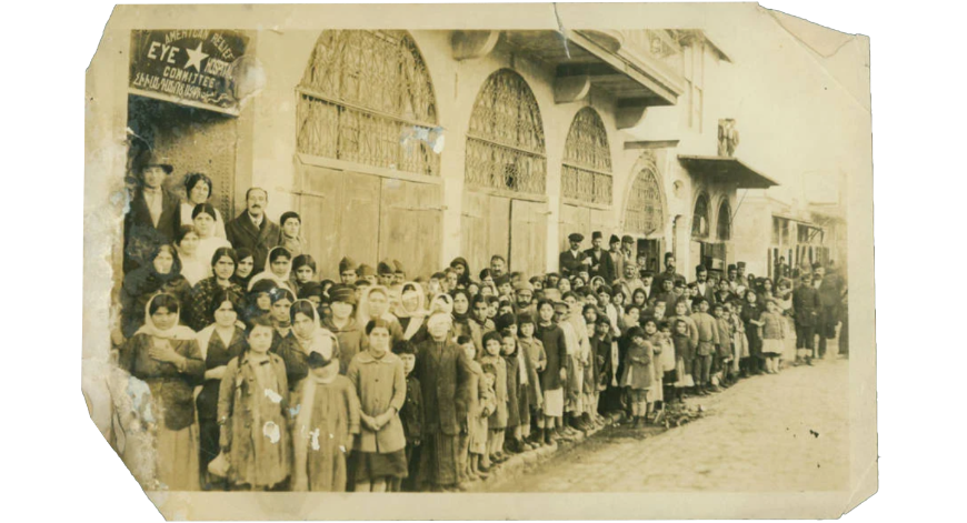 A large group of Armenian Genocide survivors, including women and children, standing outside the American Relief Eye Committee building, circa 1920s, showing the scale of humanitarian aid after The Genocide.